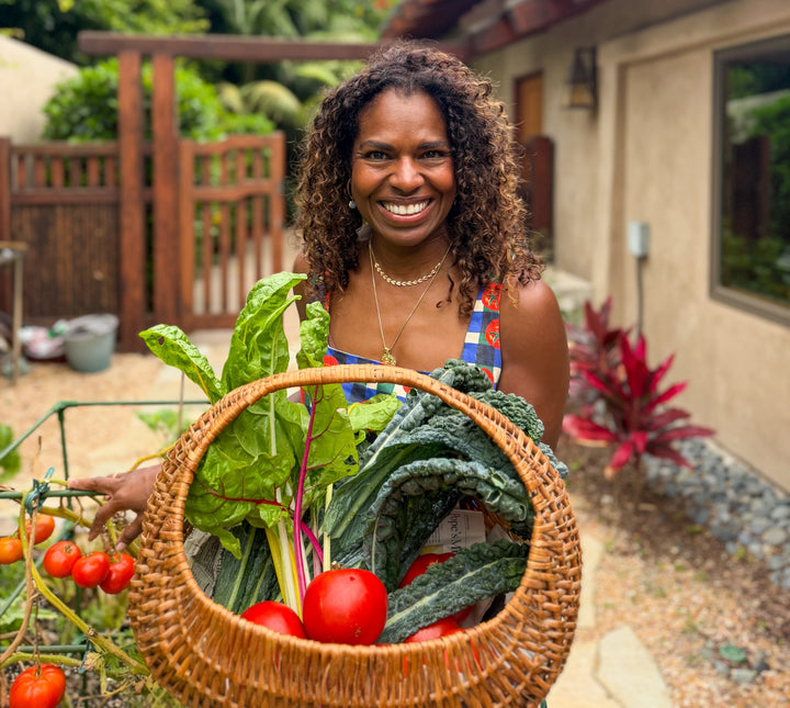 Smiling woman in a tomato print apron holding a woven basket filled with fresh garden vegetables, including Swiss chard, kale, and tomatoes, standing in a garden with a tomato vine in the foreground.