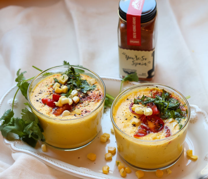 Two glass jars of sweet corn gazpacho topped with diced corn kernels, sliced cherry tomatoes, and chopped cilantro, served on a white scalloped platter next to a jar of You're So Spain Savory Spanish Spice Blend.