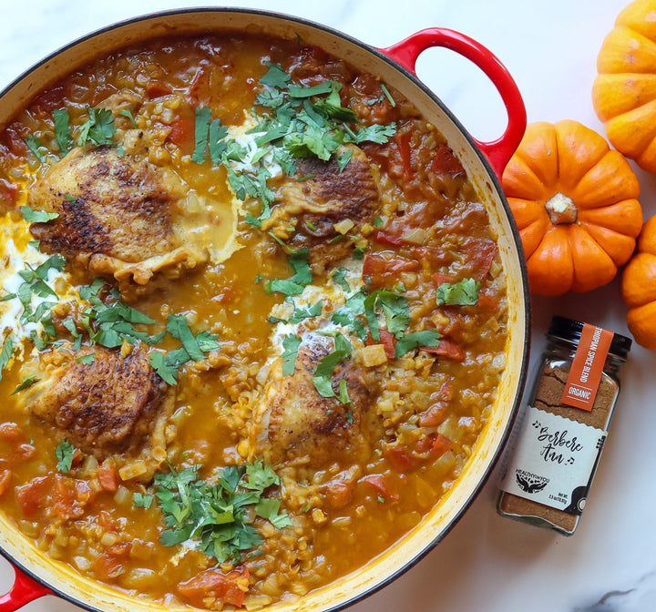 Berbere-spiced chicken thighs braised in a red lentil and pumpkin stew, garnished with cilantro and a dollop of coconut cream, served in a red Dutch oven beside a jar of Healthy On You Berbere Ann spice blend and mini pumpkins.