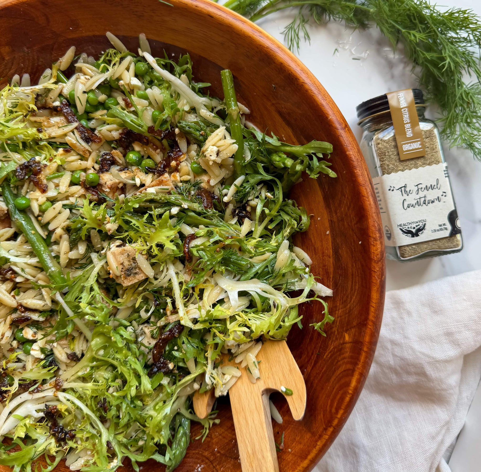 Wooden bowl with green salad and a jar of seasoning on a white surface