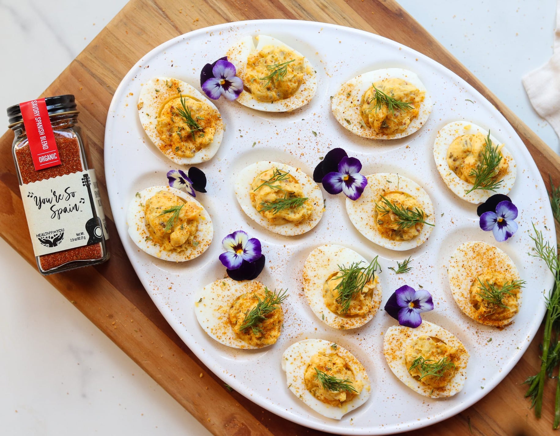Platter of deviled eggs with garnishes on a wooden board, accompanied by a container of paprika.